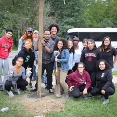A group of students pose behind a wooden post newly cemented into a hole in the ground. One student wearing gardening gloves makes a peace sign. Another student rests their hands on the handle of a shovel.