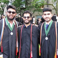 Three students in graduation gowns, with chords around draped around their necks, stand with their arms around each other. A building, trees and people are in the background.
